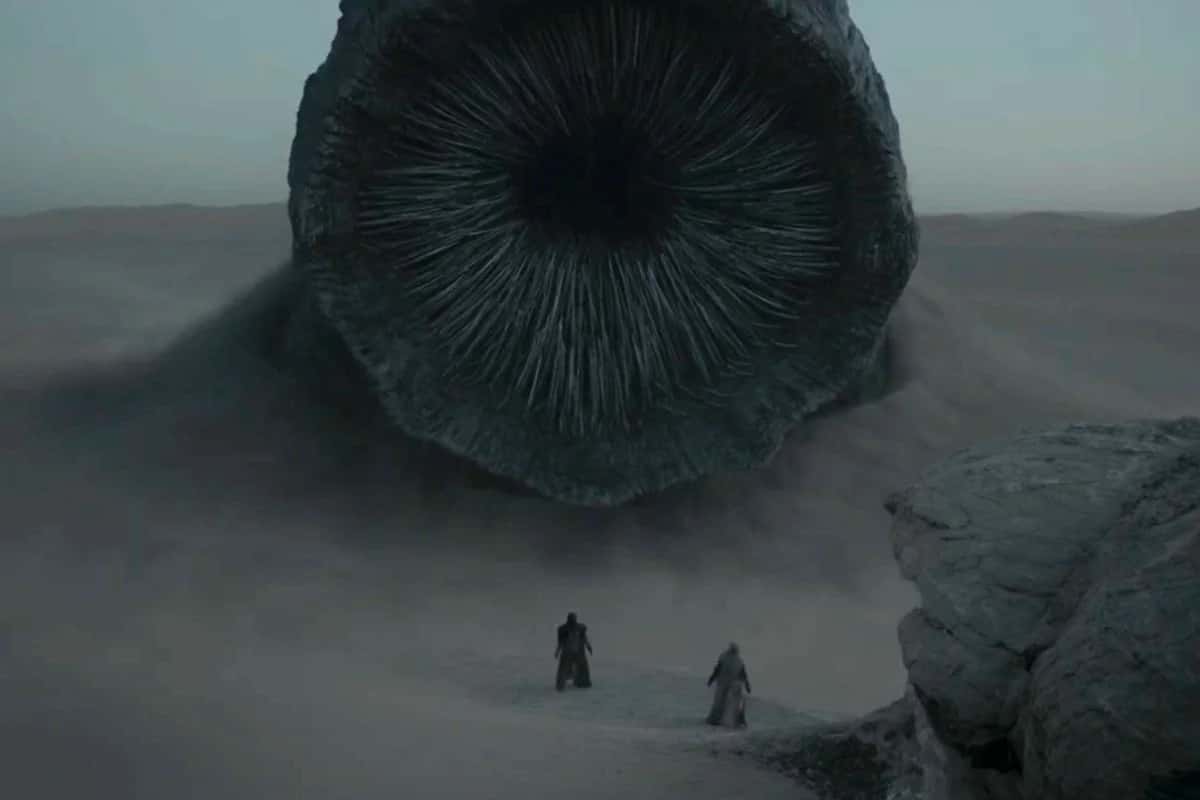 Two figures stand in a desert facing a massive worm-like creature emerging from the sand